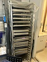 Full view of tall metal rack holding many metal baking sheets and cooling racks with plastic cover partially draped over.