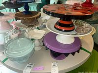 Overview of multiple ceramic and glass pedestal cake plates stacked together showing various designs and colors, including a glass cloche on a pale blue pedestal plate with a lace-like edge.