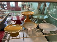 Set of six pedestal cake plates in various colors and materials displayed on white metal rack indoors.