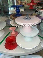 Ceramic pedestal cake plates in pastel colors including navy blue, pink with scalloped edges and decorative handle, light blue, white, and small pink. Red metal candy dish with scalloped base also visible.