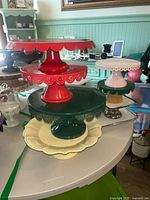 Multiple cake plates and trays displayed on a table, showing variety in colors and styles, mainly ceramic and metal with scalloped edges and raised bases.
