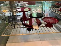 Five cake plates on a white wire shelf, including a black wire spider web cake plate and four ceramic cake plates in red and black.