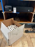 Photo of Star TSP100 thermal receipt printer on a shelf next to the cash drawer, box of receipt paper rolls on floor below.