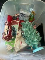 Plastic bin containing Christmas decorative items including a red and white vintage-style car, a small artificial green Christmas tree, a glittery 'Merry Christmas' item, a decorative champagne bottle, and some fabric items.