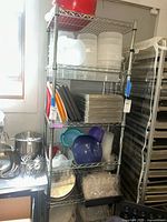 Photo shows shelving unit with stacked metal cookie sheets, clear plastic food containers, plastic bowls, and disposable cupcake trays.
