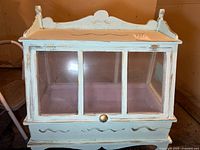 Full view of the light blue wooden display cabinet with three glass front panels, a drawer at the bottom with a round knob, and decorative carvings on the top and front.