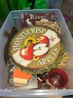 Photo showing fall-themed wall signs including 'Honeycrisp Apples' round sign and 'Rise' rectangular wooden sign, along with some crates and a small red metal bucket inside a plastic bin.