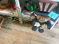 Green wire bicycle-shaped plant stand with two attached baskets next to a wooden Uncle Sam figure with red, white, and blue clothing and a pipe, placed on wooden floor.