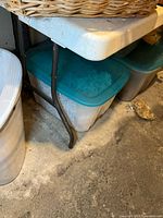 Close-up of the metal legs folding and resting on the ground beneath the table, showing the table's support structure and basement floor surface.