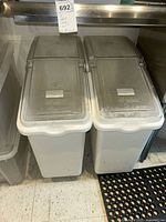 Two white rectangular plastic storage containers with clear hinged lids branded Continental Food Services Division, standing on a floor with a black anti-slip mat nearby, showing front angle and labels on lids.