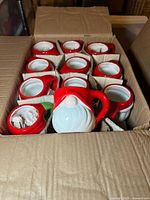 Open cardboard box showing the 23 ceramic mugs packed with cardboard separators, featuring red hat handles and white bearded gnome faces.
