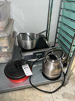 Shelf showing induction cooktop with stainless steel pot, red electric skillet with glass lid, kettle on base station