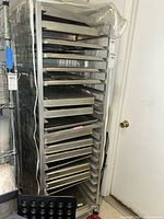 Metal industrial kitchen rack holding multiple metal baking trays in an indoor room corner, some trays have wire mesh design.