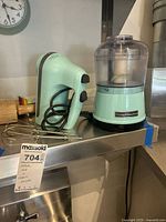 KitchenAid hand mixer and mini food processor displayed side by side on a stainless steel counter, showing the beaters and cords.