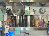 Two metal containers on a metal shelf holding white and green silicone brushes, spatulas, whisk. Metal strainers and other utensils visible alongside. Plastic container to side.