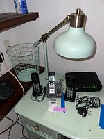 Image showing metal desk lamp with adjustable arm, three black Panasonic cordless phones, wire waste basket, and Arris modem on a small white table.