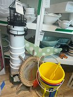 White decorative lighthouse, nautical ship wheel mirror, and plastic buckets with attached shovels shown on wooden floor and against shelving with housewares.