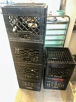 Six stacked black plastic milk crates featuring lattice design and integrated handles for carrying, shown indoors with other household items