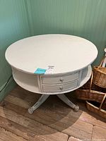 Front angle view of round white painted wood occasional table showing single drawer with two knobs and open shelf underneath, pedestal legs with carved feet, some signs of damage and wear.
