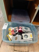 Photo showing a clear plastic bin filled with various paper punches of multiple colors and styles placed on a wooden floor inside shelving space.