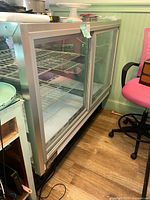 Metal and glass refrigerated display cabinet showing front and side view with sliding glass doors and wire shelves inside on wooden floor near pink chair.