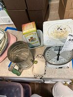 Photo showing metal sifter, Droste's Cocoa tin, scale and part of the bowl and tray on a table surface.