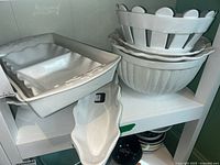 Photo showing stacked ceramic bowls and several serving dishes on a white shelf. Pieces are white with decorative edges and ribbed surfaces on mixing bowls.