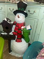 Front view of snowman figure wearing black top hat with holly, red and green scarf, holding green stocking and birdhouse with Welcome sign.