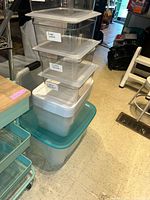 Overall view of stacked clear plastic storage bins with lids, each labeled, placed on and inside a large blue plastic storage bin in a cluttered room.