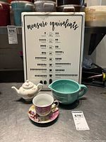 Lot items including measure equivalents sign, teapot, teacup with saucer, and measuring cup arranged on metal surface