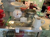Photo showing vintage metal kitchen scale, white ceramic pitcher, metal cake pans, gold-tone Welcome sign, and small business card holder placed on a glass shelf.