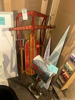 Photo showing vintage red wood and metal sled labeled 'Rocket Steering Astro' with metal runners and wooden slats, leaning against wall.