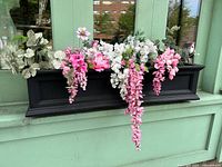 Front view of black plastic window box planter filled with cascading faux flowers in pink, white, and light green tones displayed on a green window ledge.