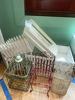 White wooden picket fence style boxes, white angled wooden box with handle, metal birdcage decor, pink metal wire chair, cream metal planter box with scroll handles, all shown on wooden floor.