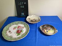 Three antique decorative plates displayed on a blue cloth, showing floral and village scenes from Bavaria and Germany.