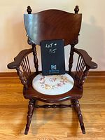 Front view of antique dark wood rocking chair showing padded floral embroidered seat, high solid wood backrest with finials, and spindle arm supports, on wood floor.