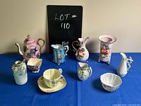 Photo of antique and vintage porcelain and ceramic items including teapot, pitchers, cups, and vases arranged on a blue cloth against a plain wall. Items show decorative hand-painted details, one marked Lefton China.