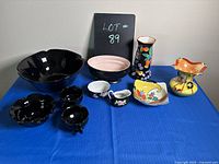Full view of the eclectic mix of black and colorful dishes on a blue surface with black backdrop sign reading 'LOT 89'. Items include various bowls, creamers, a pitcher, and decorative pieces.