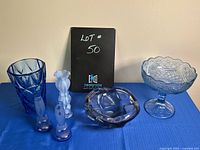 Photo showing the entire lot of blue glass pieces on a blue cloth background, clearly displaying the vase, compote bowl, decorative bowl, two candle holders, and textured glass piece.