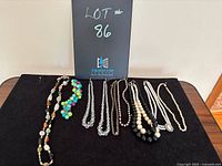 View of 11 costume jewelry beaded necklaces arranged on a black cloth, showing variety of styles including colorful beads, faux pearls, and crystal-like beads.