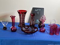 Nine pieces of cranberry and red glass items displayed on blue cloth, including vases, bowl, and drinking glasses