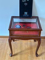 Front angled view of wooden display case with glass top and sides, showing red lining and stone brooches/pendants inside.