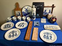 Top view of a blue cloth covered table displaying various Dutch themed ceramic plates, mugs, wooden plaques, framed pictures, and vases arranged neatly.