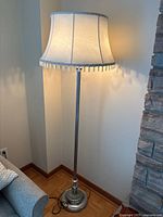 Floor lamp with light-colored drum shade with tassels, metal stem, and round base showing metal and stone-like textures, standing in corner of room next to couch and stone wall.