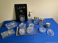 Photo of a collection of clear glass vanity items including a soap dispenser with metal pump, drinking goblet, rectangular trays, multiple glass containers with lids, and ribbed oval jewelry holders.