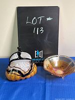 Glass purse-shaped vase and carnivale glass candy dish on blue cloth with black backdrop labeled Lot 113