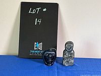 Two indigenous stone carvings on blue cloth background, one a face and one a seated man figure