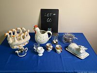 Overall view of all kitchen items on blue fabric - duck planter, rooster pitcher, cow butter dish, egg cups, small teapot, hourglass timer, glass teapot.