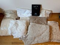 Group of nine lace tablecloths in white, beige, and off-white colors placed on a wooden floor. Some are folded and some packaged in plastic.