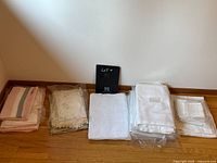 Lot of five groups of folded linen tablecloths and napkins arranged on a hardwood floor against a wall. Includes pastel striped fabric, embroidered fabric, lace fabric, plain white linens, and smaller white linens still in plastic.
